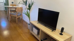 Ein Wohnzimmer mit Fernseher auf einem Holzständer, einem Esstisch mit Stühlen, einer Topfpflanze und einer Fernbedienung auf dem Ständer.