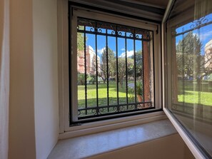 Ein Fenster mit einem Metallgitter, Blick auf eine grüne Rasenfläche und ein Gebäude im Hintergrund.