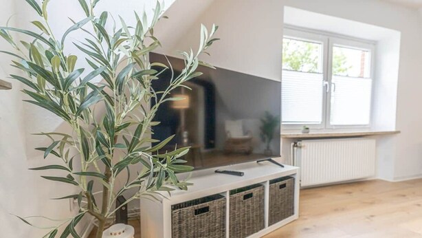 A room with a TV on a white stand, wicker baskets, a plant, and a lantern. - Bright & Cozy Apartment with Harbor View in Varel (Varel)