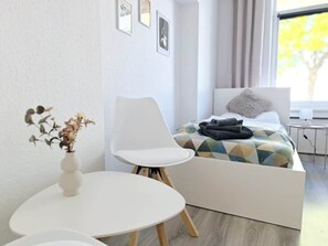 Un dormitorio moderno con una cama, una silla blanca, una mesita y un florero con flores secas.