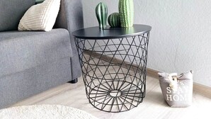 Ett runt metallbord med ett geometriskt mönster, en grå soffa med en kudde och en liten kaktusplanta på bordet.