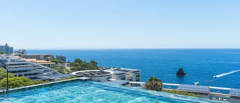 Piscina infinita en la azotea con vistas a una ciudad costera, edificios y el mar.