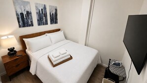 Habitación de hotel con cama, mesita de noche, lámpara, televisión y plancha mural.