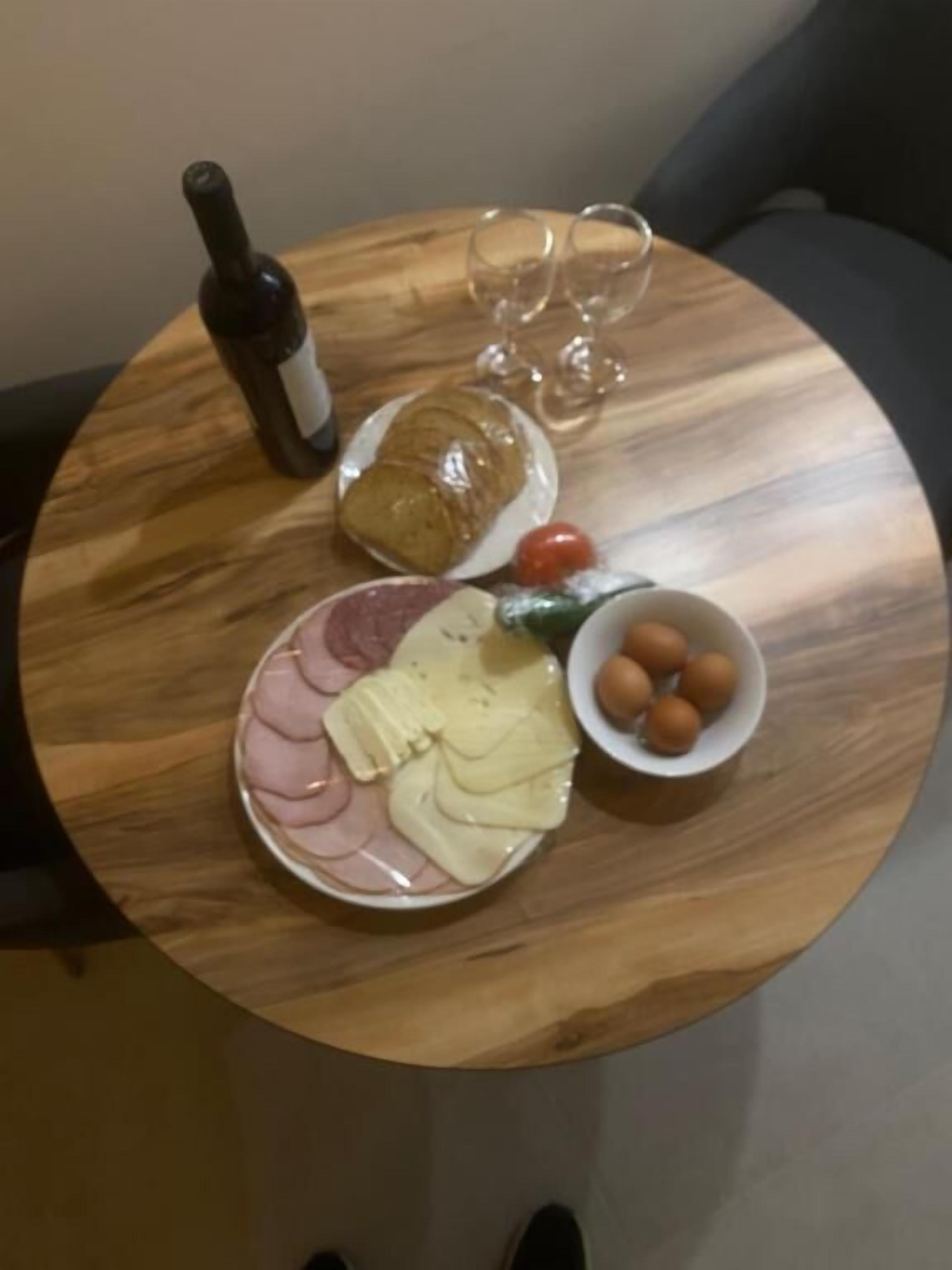 Een ronde houten tafel met een fles wijn, twee glazen, een bord met kaas en ham, een broodje, een klein schaaltje olijven en een tomaat.