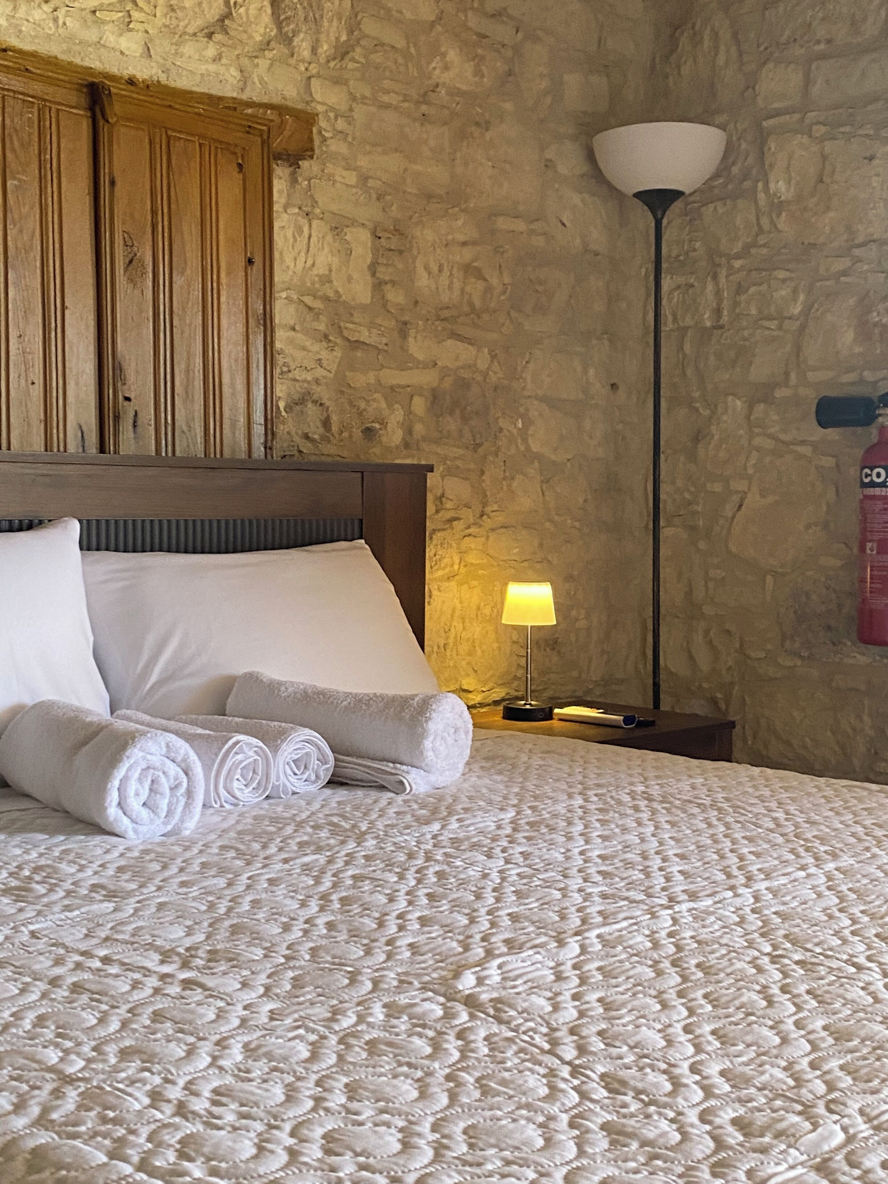 Una cama bien hecha con toallas enrolladas, una lámpara de noche y una pared de piedra.