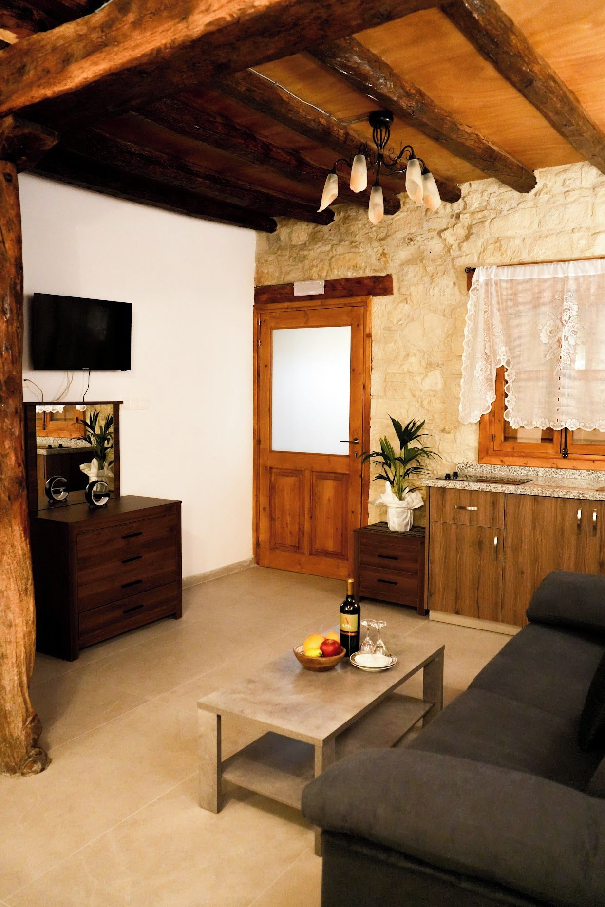 A rustic living room with a wooden ceiling, a television mounted on the wall, a wooden door, a sofa, a coffee table with fruit and a bottle, and a kitchen area with a window.