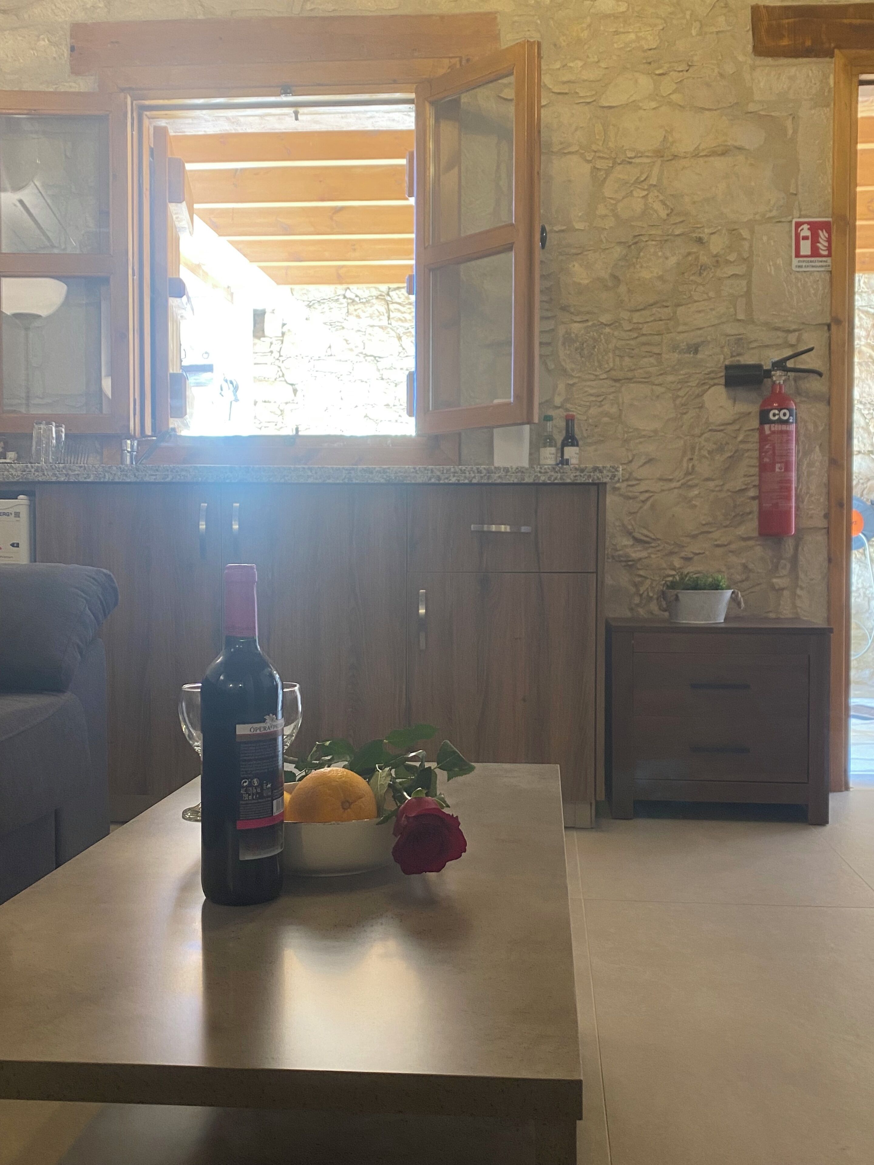 A kitchen with wooden cabinets, a countertop with a wine bottle, a bowl with an orange and a rose, and a fire extinguisher on the wall.