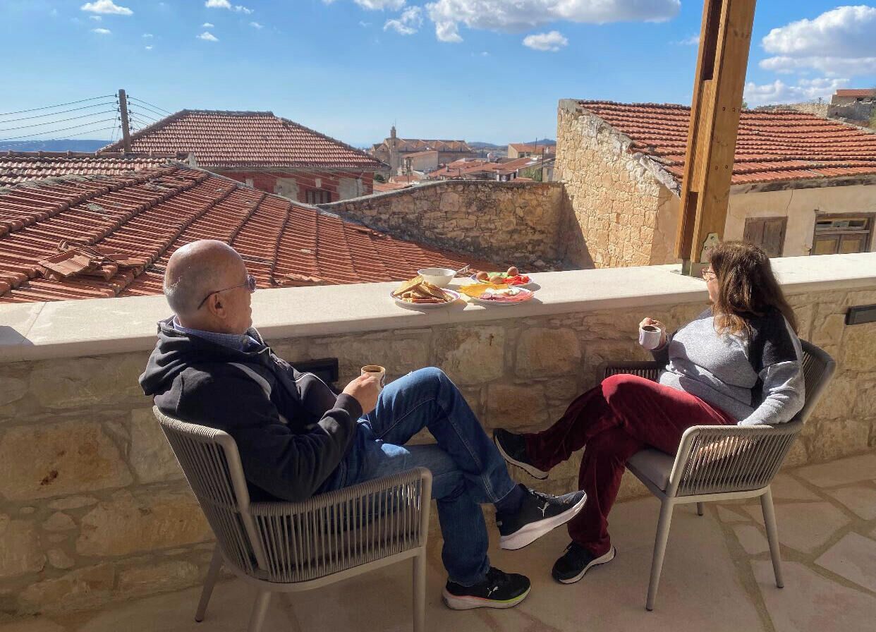 Dos personas sentadas en sillas al aire libre, con una mesa entre ellas, contemplando una vista urbana de tejados rojos.