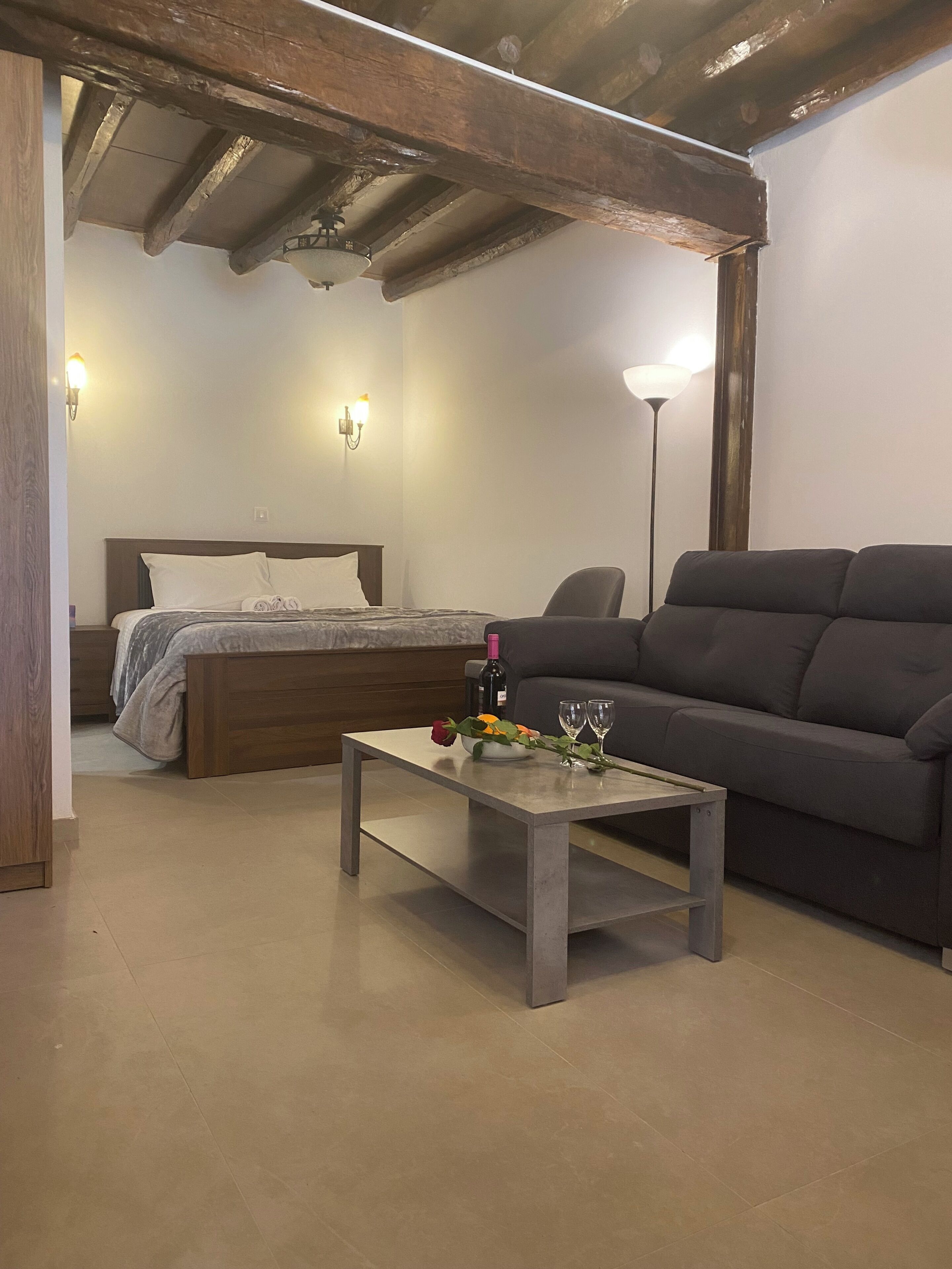 A room with a bed, a sofa, a table with a rose and wine glasses, and a lamp.