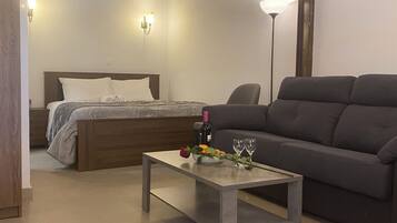 A room with a bed, a sofa, a table with a rose and wine glasses, and a lamp.