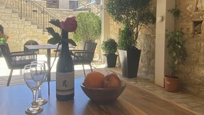 A wooden table with a wine bottle, glasses, and a bowl of fruit, set against a stone wall and a view of a building with a staircase. - Vouni Calais  (Vouni)