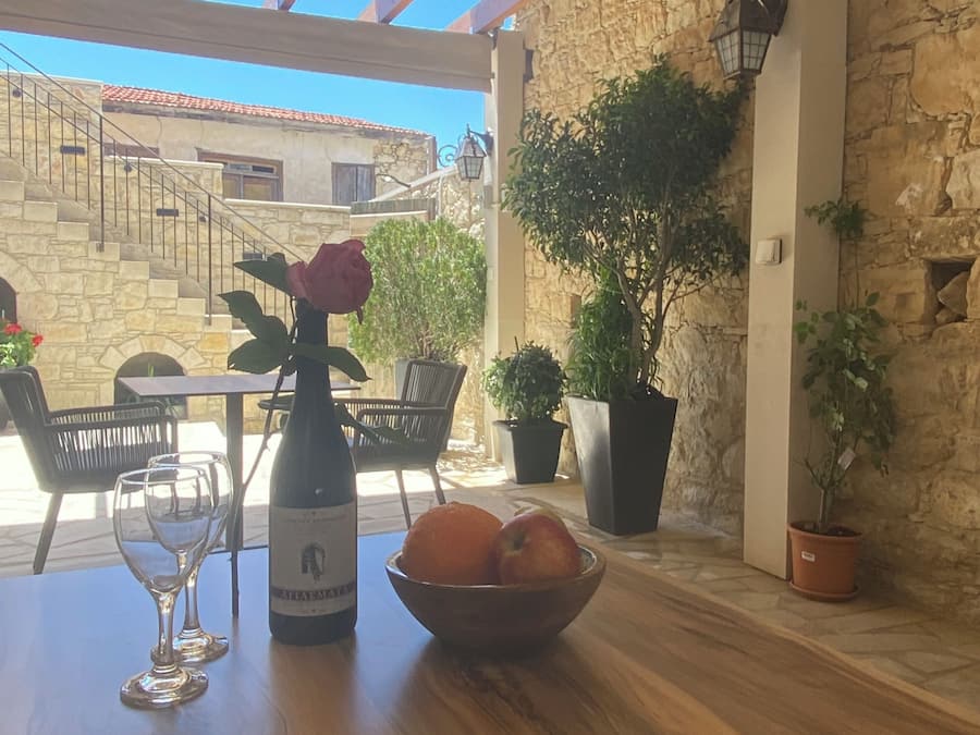 Una mesa de madera con una botella de vino, copas y un bol de fruta, frente a una pared de piedra y vistas de un edificio con escaleras.