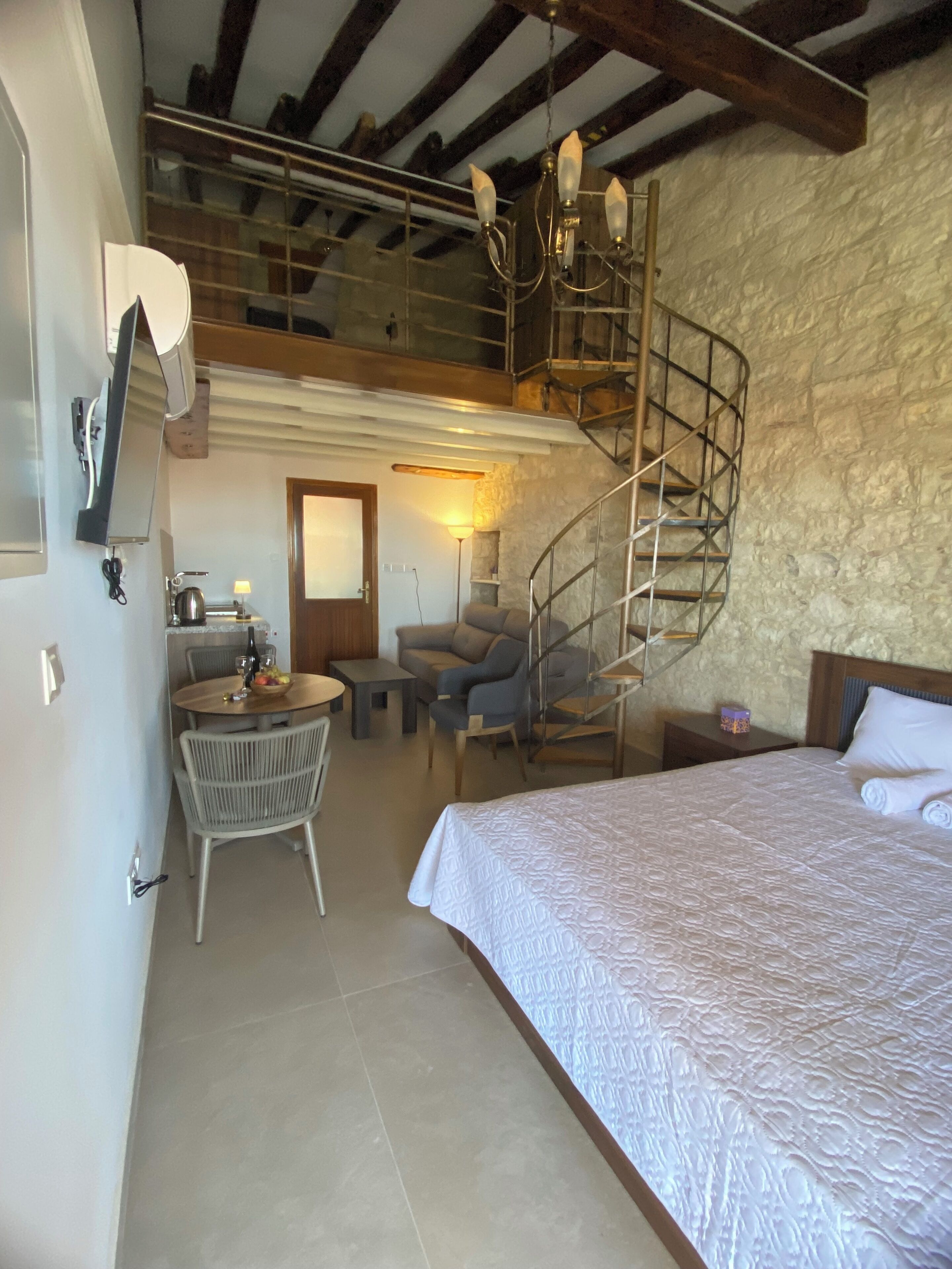 A bedroom with a large bed, a dining area with a table and chairs, a staircase, and a stone wall.