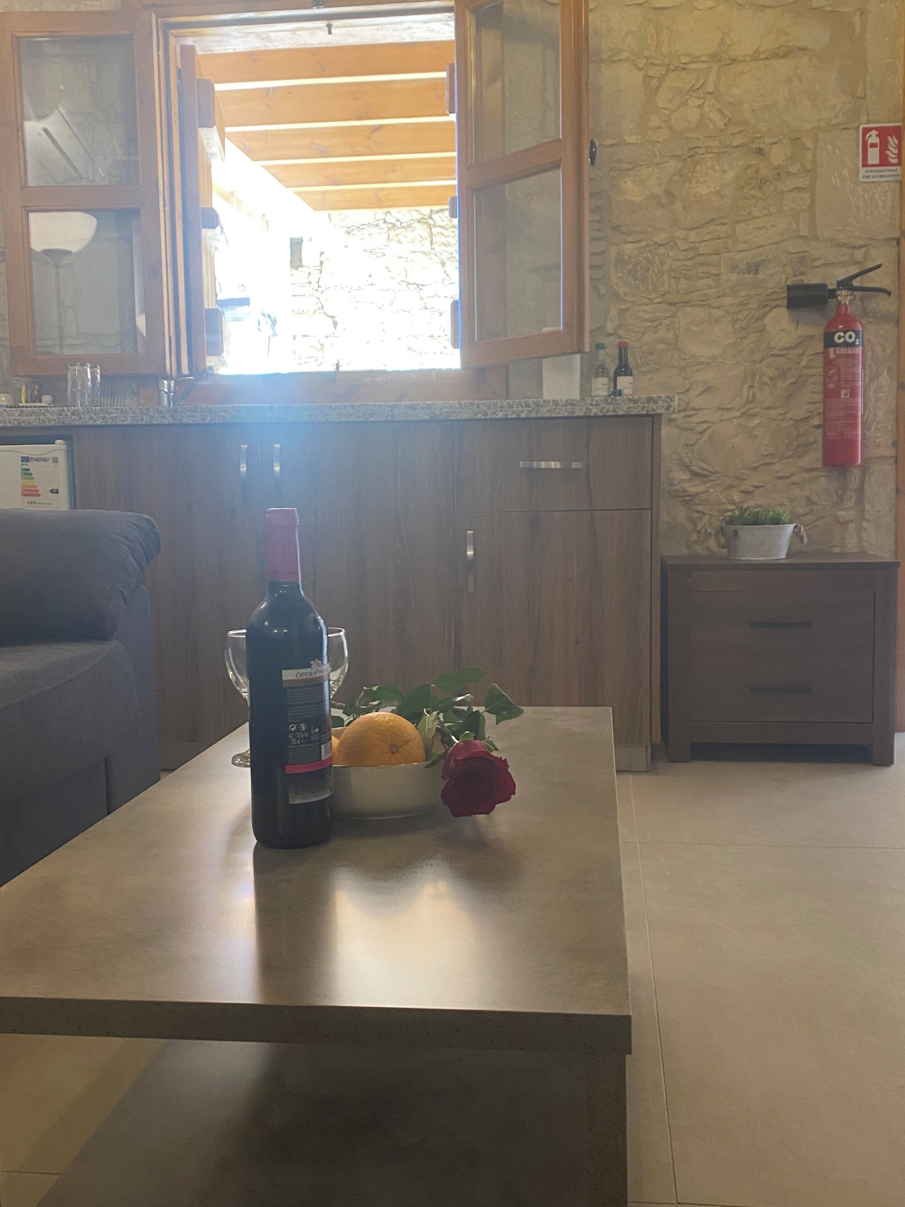 Una cocina con armarios de madera, una encimera, una botella de vino, un bol con una naranja y una rosa, y un extintor en la pared.