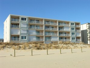 Hotel de vários andares com varandas, situado numa praia de areia com dunas ao fundo.