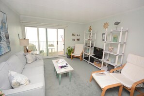 A living room with a white sofa, a coffee table, and a TV stand with shelves. - Kitschy 1 Bedroom Oceanfront Condo with Free WiFi, Coastal Decor, and a Breathtaking View Located Uptown and Mere Steps from the Beach! (Ocean City)