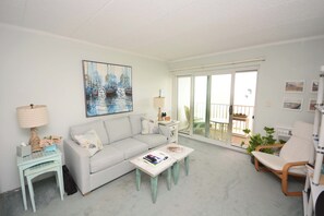 A living room with a sofa, coffee table, and chairs, a painting on the wall, and a balcony with plants. - Kitschy 1 Bedroom Oceanfront Condo with Free WiFi, Coastal Decor, and a Breathtaking View Located Uptown and Mere Steps from the Beach! (Ocean City)