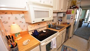 Una cocina con armarios blancos, microondas, estufa y pileta. Hay una tetera azul sobre la estufa, una lámpara en la encimera y un tapete con estampado de peces en el piso.