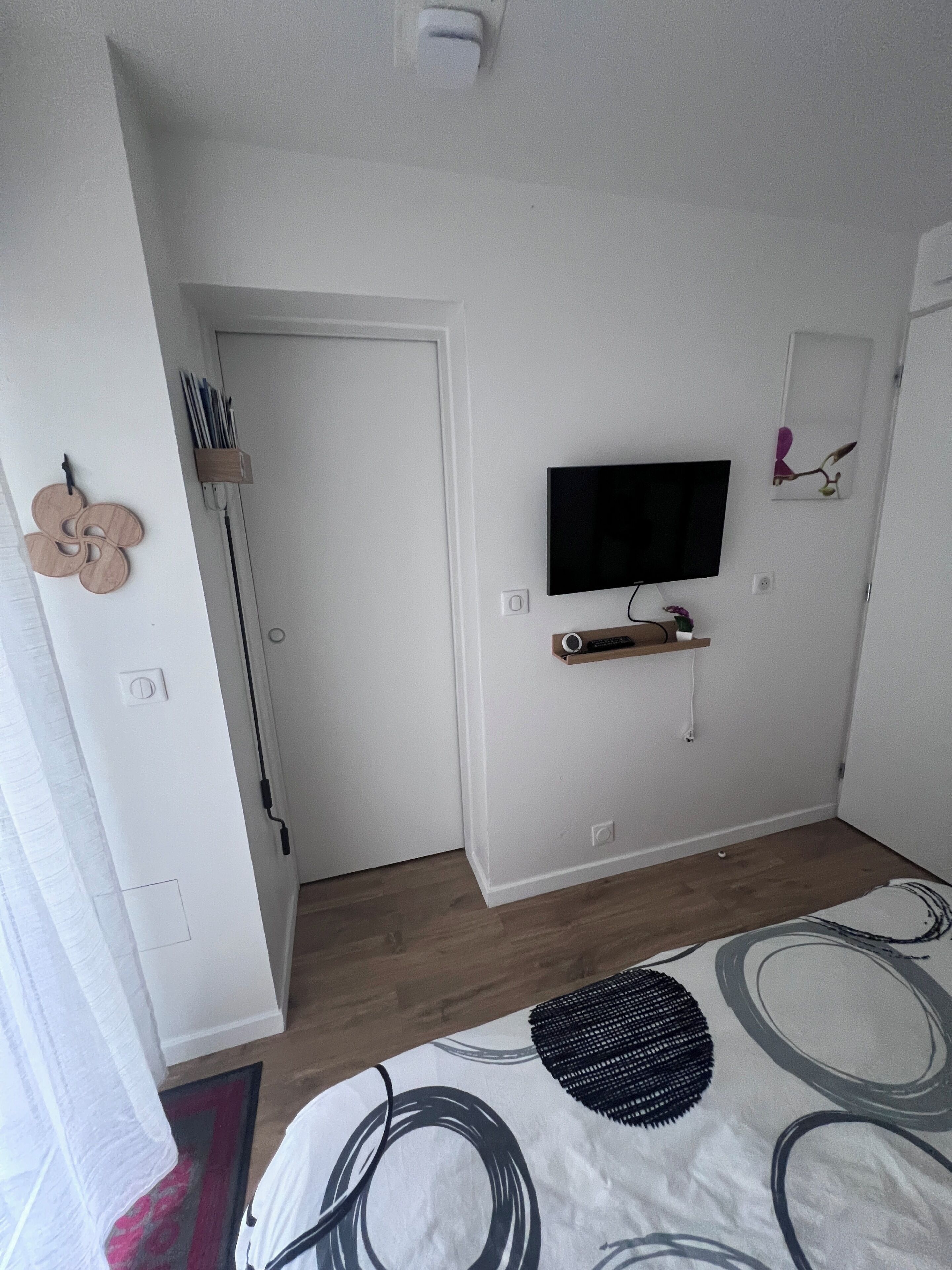 Un dormitorio con cama, televisor montado en la pared, una puerta y un estante pequeño con un control remoto.