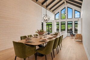 A dining room with a long table set for six, surrounded by green upholstered chairs, a chandelier, and large windows offering a view of greenery. - Tremblant Prestige - Verbier 10-203 (Mont-Tremblant)