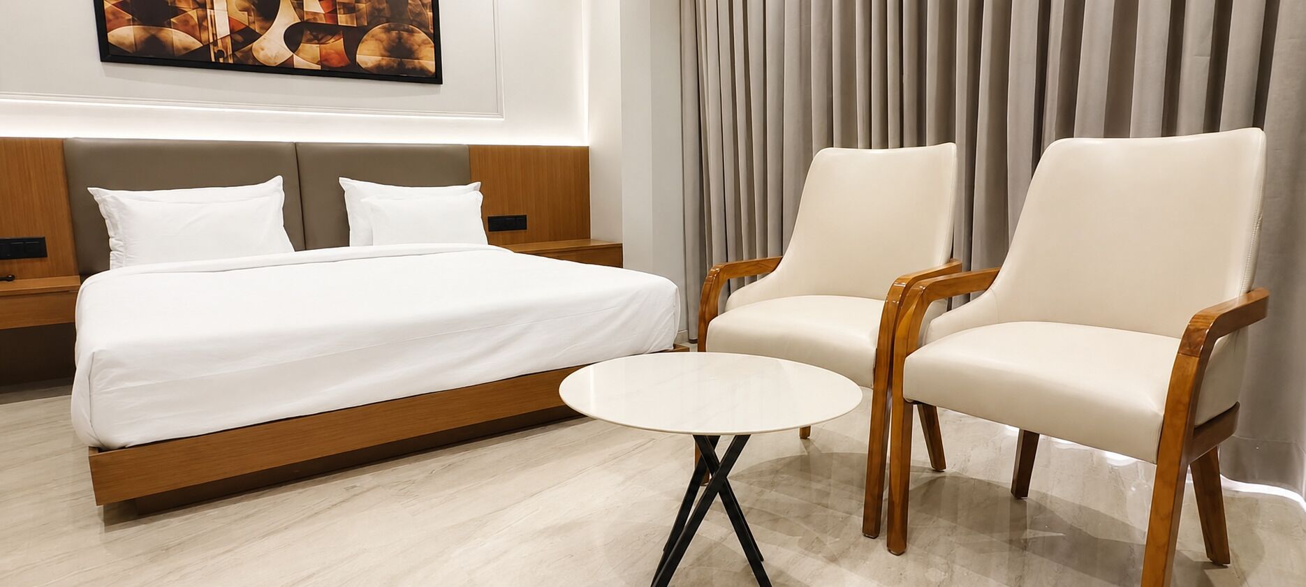 A hotel room with a bed, two armchairs, a small round table, a painting on the wall, and a curtain.