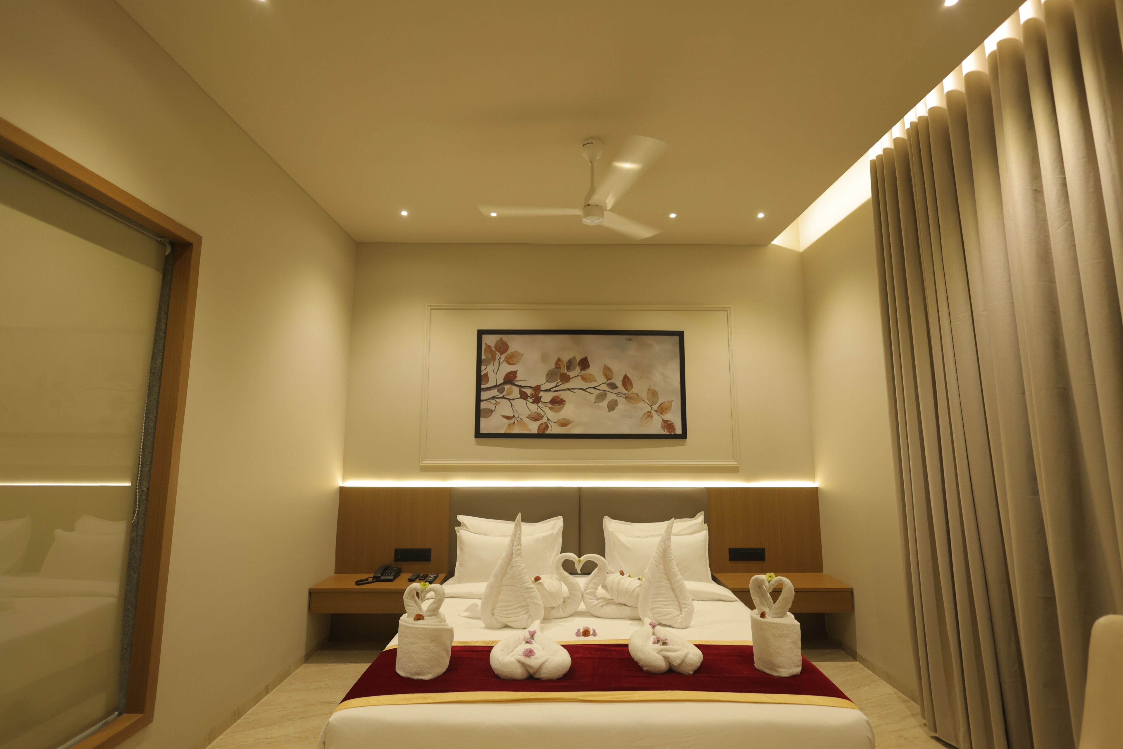 Una habitación de hotel con una cama grande, un ventilador de techo, una obra de arte enmarcada sobre la cama y una ventana con persianas.
