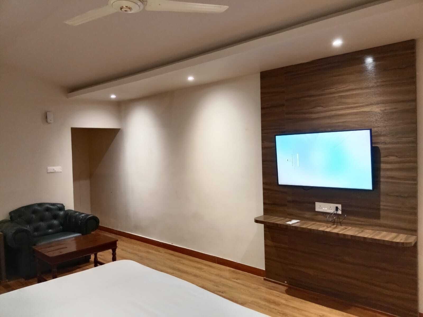 A modern hotel room with a flat-screen TV mounted on a wooden wall, a leather sofa, a wooden coffee table, and a bed with a white mattress.
