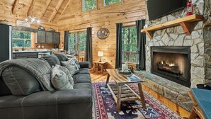 Ein Blockhaus-Interieur mit Kamin aus Stein, einer Couch mit Kissen, einem Essbereich und einer Küche.