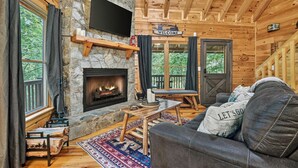 Ein Cabin-Innenraum mit Kamin aus Stein, einem darüber montierten Fernseher, einem 'WELCOME'-Schild, einer Couch mit Kissen mit der Aufschrift 'LET'S GO' und einem durch die Fenster sichtbaren Holzboden.