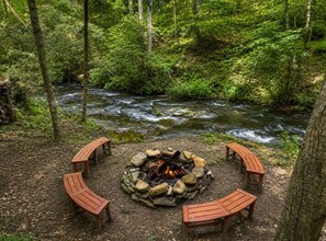 Ein kreisrunder Feuerkorb mit umlaufenden Bänken, eingebettet in einen Wald, im Hintergrund fließt ein Fluss.