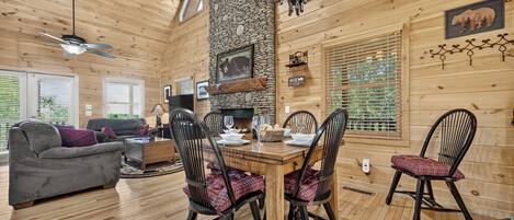 A wooden cabin interior with a dining area, fireplace, and a living room with a sofa and TV.