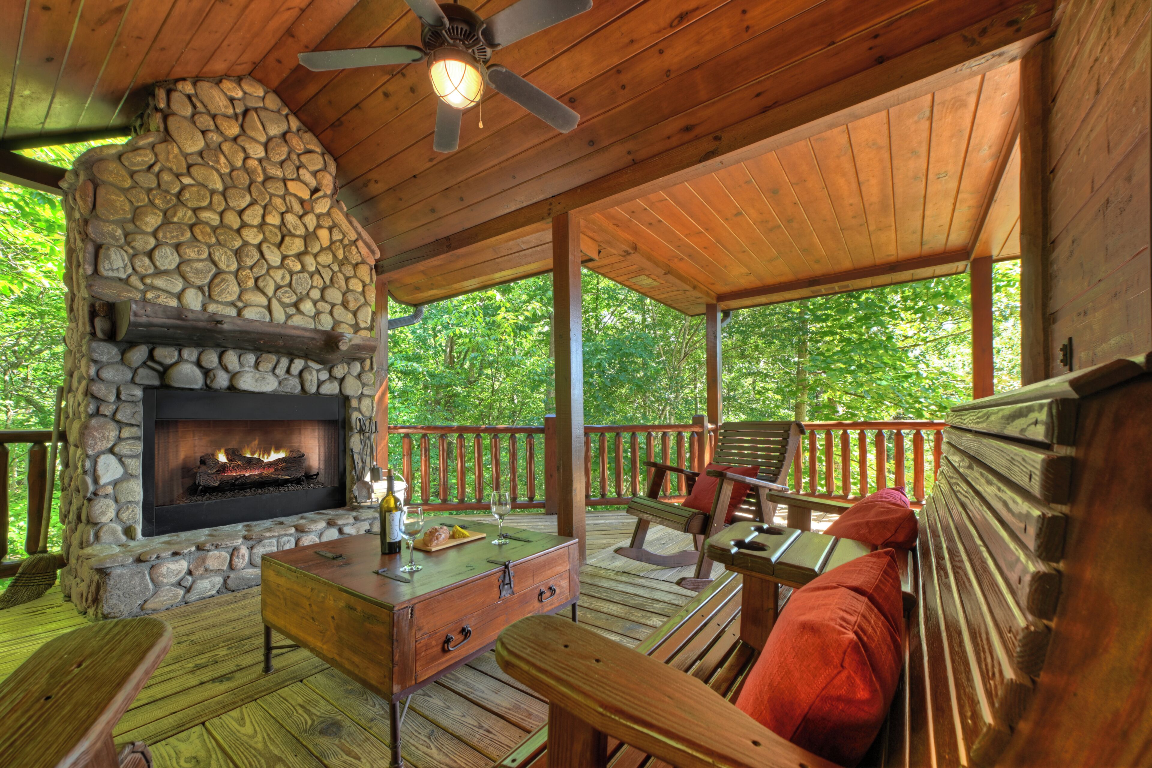 A covered outdoor deck with a stone fireplace, wooden furniture, and a view of the surrounding greenery.