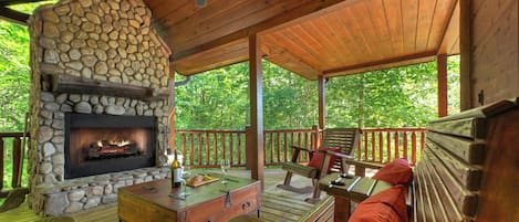 A covered outdoor deck with a stone fireplace, wooden furniture, and a view of the surrounding greenery.