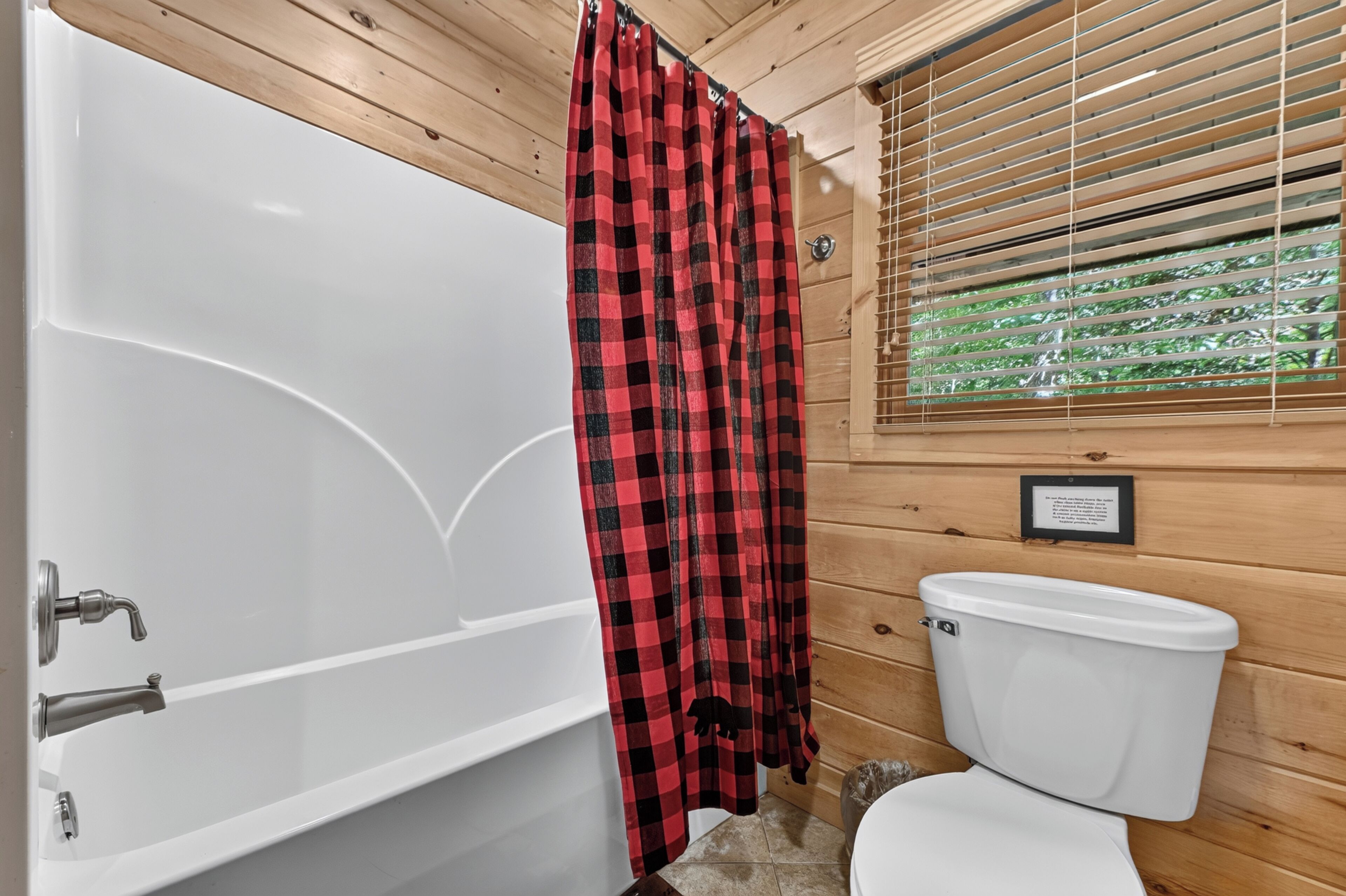 A bathroom with a white bathtub, a red and black checkered shower curtain, a white toilet, and wooden walls.