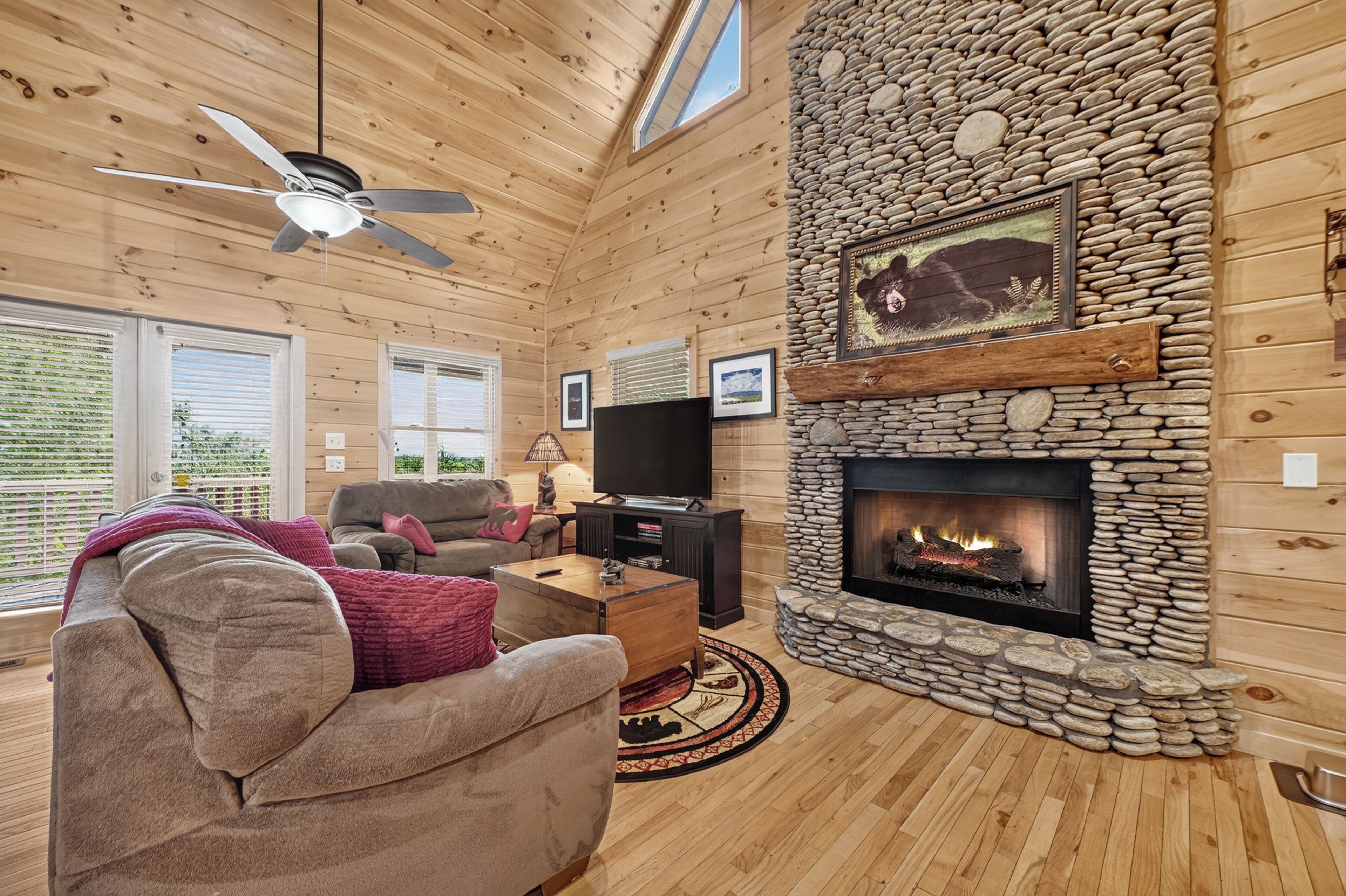 A cozy cabin interior with a stone fireplace, wooden walls, a ceiling fan, and a television.