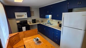 A compact kitchen with dark blue cabinets, a white microwave, a white refrigerator, and a wooden dining table with a bowl. - Birch Bungalow - Unit D a cozy 2-bedroom retreat in charming Sault Ste. Marie! (Sault Ste. Marie)
