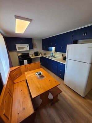 Une cuisine compacte avec des meubles de couleur bleu foncé, un micro-ondes blanc, un réfrigérateur blanc et une table de salle à manger en bois sur laquelle se trouve un saladier.