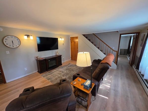 A living room with a flat-screen TV, a clock, a sofa, a coffee table, and a staircase.