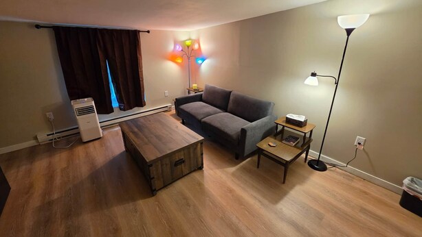 A living room with a sofa, a wooden coffee table, a standing lamp, and a heater. - Birch Bungalow - Unit D a cozy 2-bedroom retreat in charming Sault Ste. Marie! (Sault Ste. Marie)