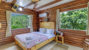 Quarto de cabana de madeira com cama, cortinas, ventilador de teto e janela com vista para vegetação.