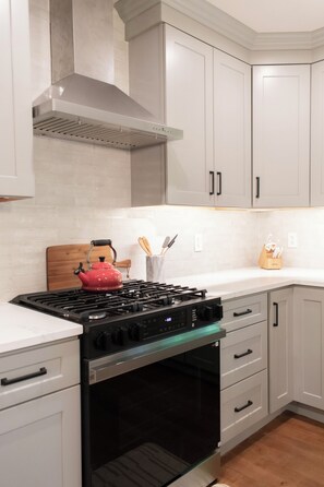 Una cocina con armarios blancos, una campana extractora de acero inoxidable, un hervidor rojo y una tabla de cortar de madera.