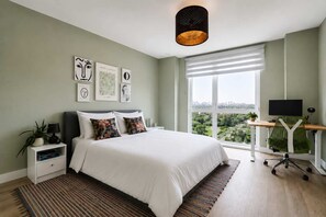 A bedroom with a bed, bedside tables, a desk with a chair, a large window, and artwork on the wall. - The London Horizon with Air-con (London)