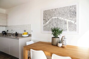 A modern kitchen with a white countertop, a wooden dining table, and a large cityscape artwork on the wall. - East London Views - Modern Apartment (Greater London)