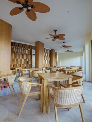 A spacious indoor area with multiple wooden tables and chairs, ceiling fans, and a wooden bookshelf. - Kinan Sentral Maldives (Fulidhoo)