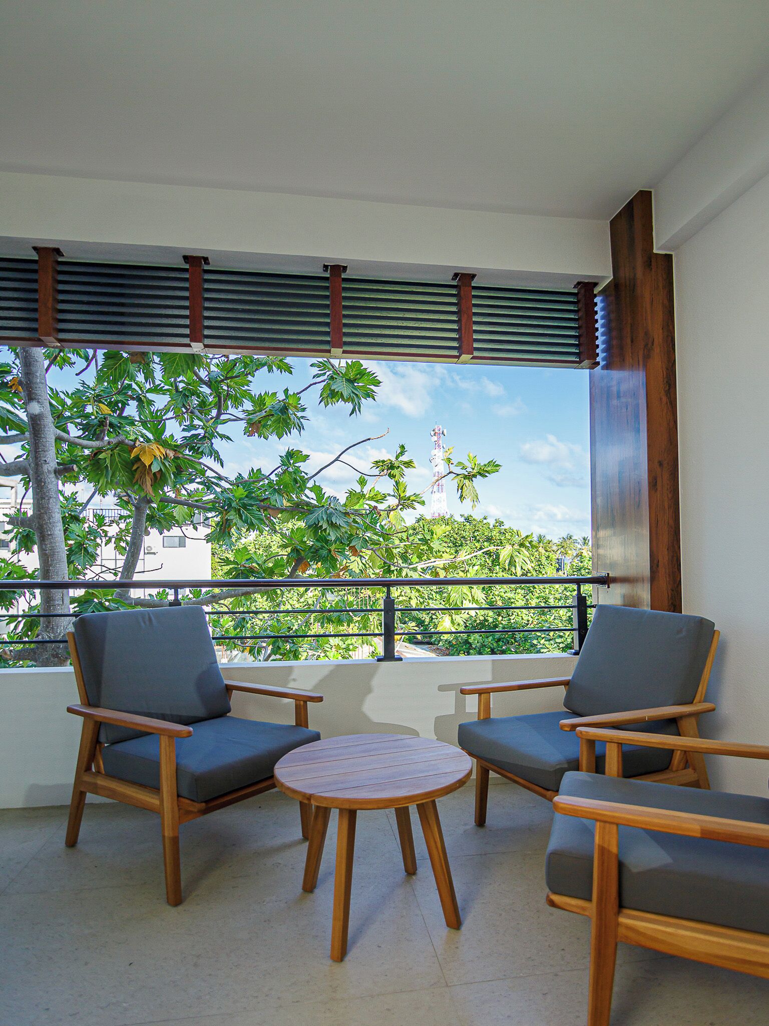 A balcony with two wooden chairs and a small round table, overlooking a lush green landscape with trees and a clear sky.