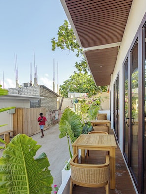 Un patio con mesas y sillas de madera, una planta grande y frondosa, y vista a una zona residencial con un muro de ladrillo y una persona caminando.