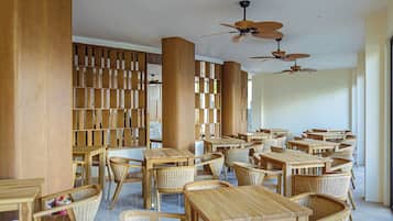 A spacious indoor area with multiple wooden tables and chairs, ceiling fans, and a large bookshelf.
