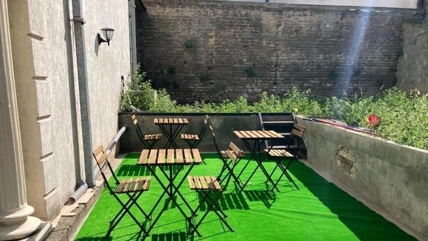 Un pati exterior amb gespa artificial verd, taules i cadires de fusta i un mur de maó al fons.