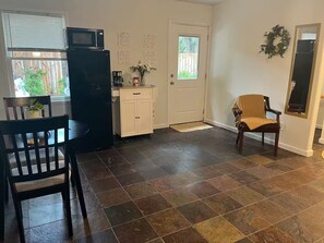 A kitchen with a refrigerator, microwave, and cabinets. There is a dining table with chairs and a small potted plant. A door with a doormat is visible.