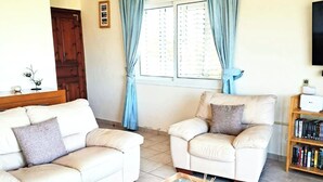 A living room with two white leather sofas, a glass coffee table, a wooden bookshelf, and a window with blue curtains. - Villa Koryfi - Argaka - 3 Bedroom villa minutes walk from the beach (6 guests) (Argaka)